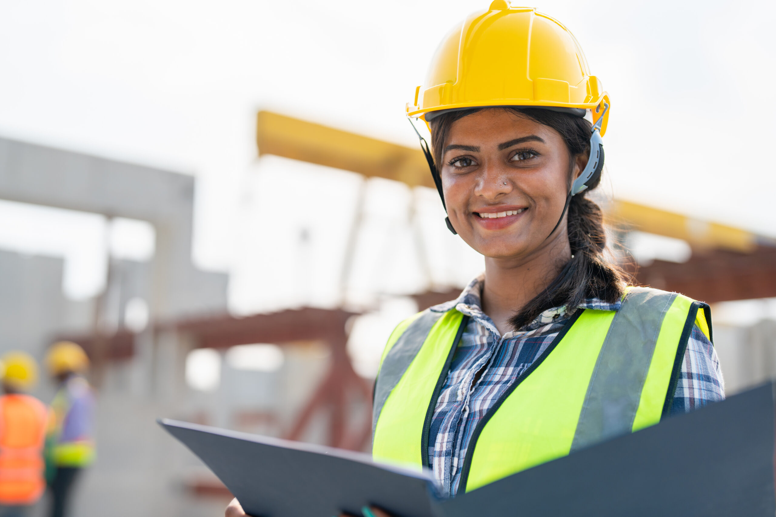 Asian woman engineer holding document smiling at construction site. Confident female Indian wearing protective helmet and vest working in factory marking precast concrete wall for real estate housing.